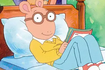 Arthur the cartoon sitting on a bed reading a book